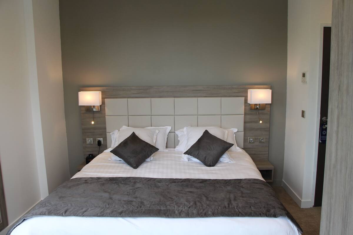 A neatly made bed with a grey blanket, two dark pillows, and a headboard with wall-mounted lamps.