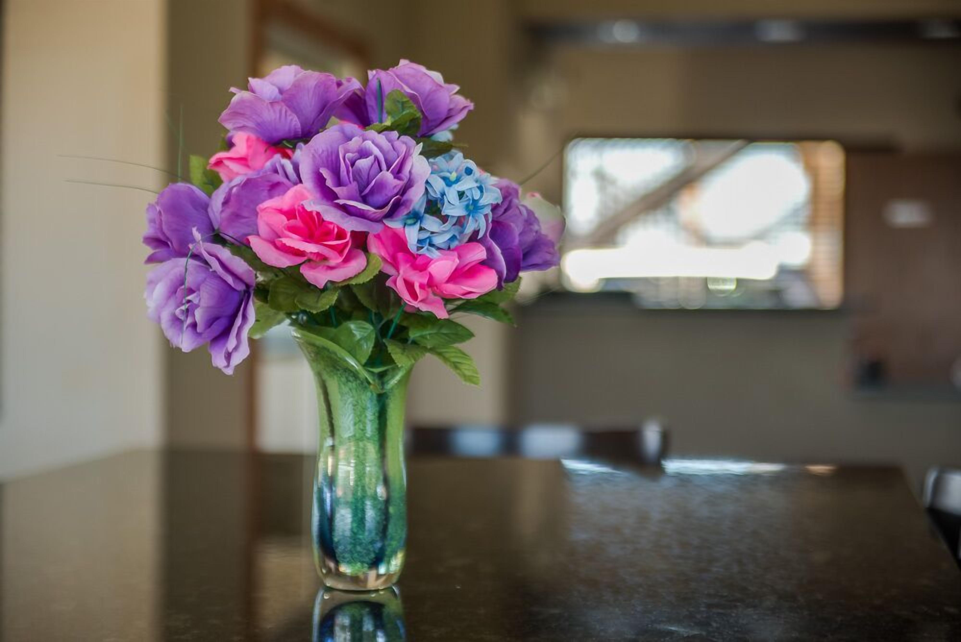 A vase with a bouquet of purple, pink, and blue flowers on a reflective surface.