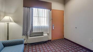 A hotel room with a patterned floor, a blue sofa, a standing lamp, a window with curtains, and a door.