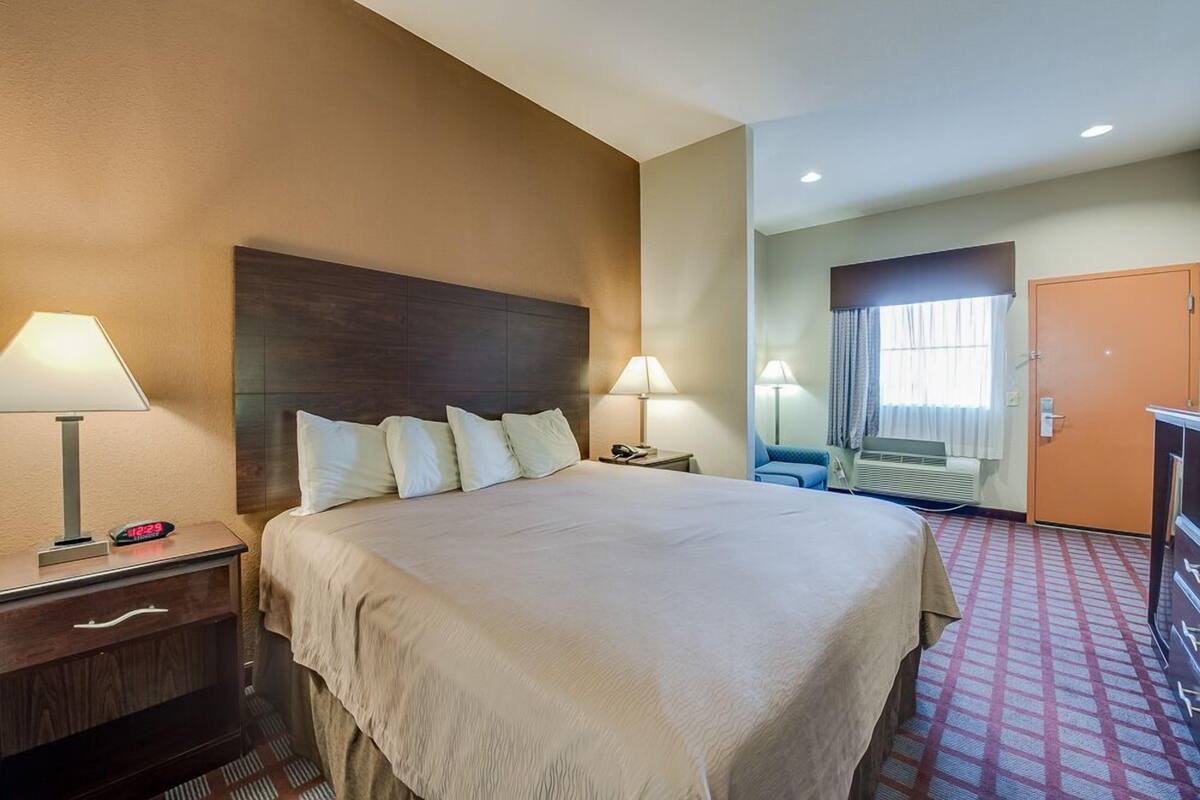 A hotel room with a large bed, two bedside tables with lamps, a clock, a desk with a chair, and a window with curtains.