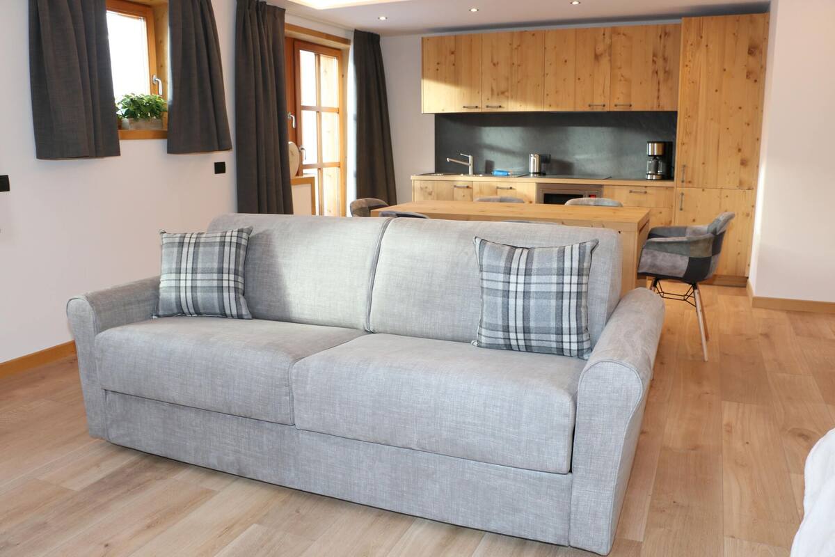 Ein modernes Wohnzimmer mit einem grauen Sofa, zwei karierten Kissen, einem Holzesstisch und einer Küchenzeile mit Holzschränken und einer schwarzen Arbeitsplatte.