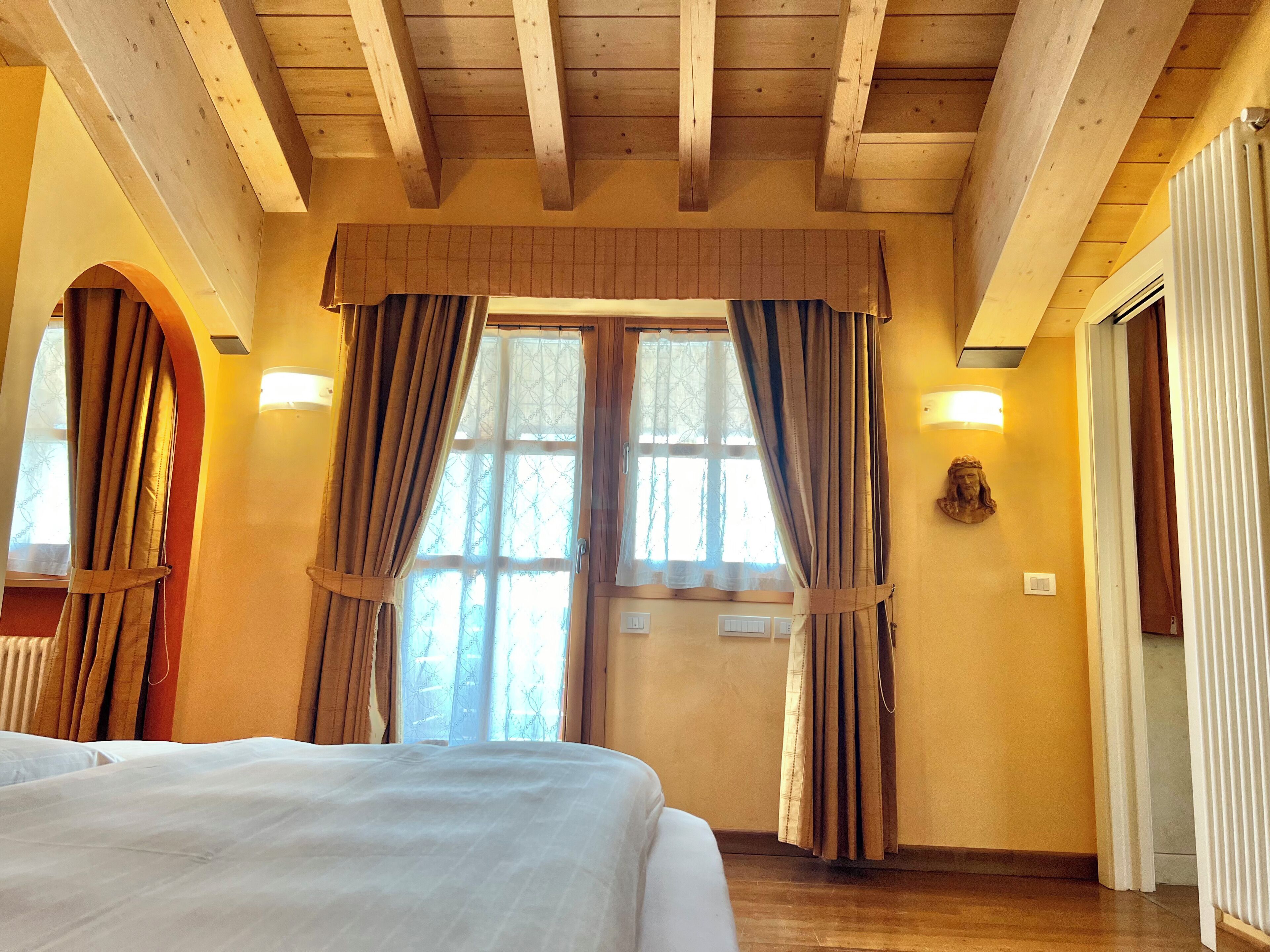 A bedroom with a wooden ceiling, a large bed, two windows with curtains, and a wall-mounted light fixture.