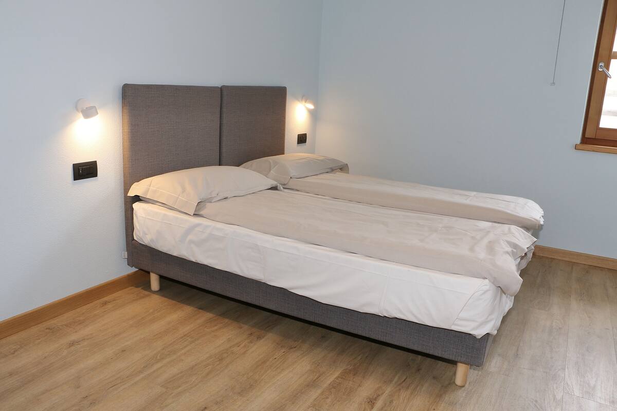 Ein Doppelbett mit grauem Kopfteil und zwei Nachttischen, an denen jeweils eine Wandleuchte angebracht ist.