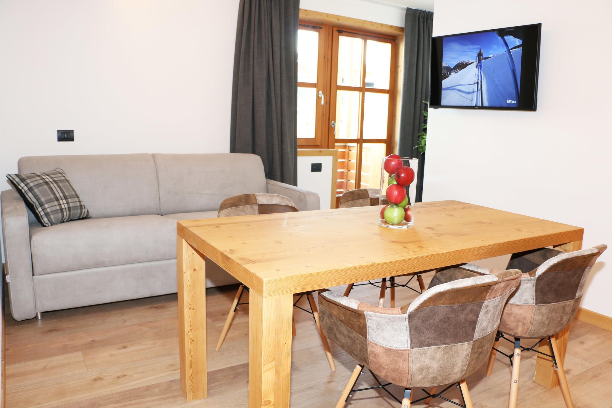 A living room with a wooden dining table, four chairs, a sofa, and a television on the wall.