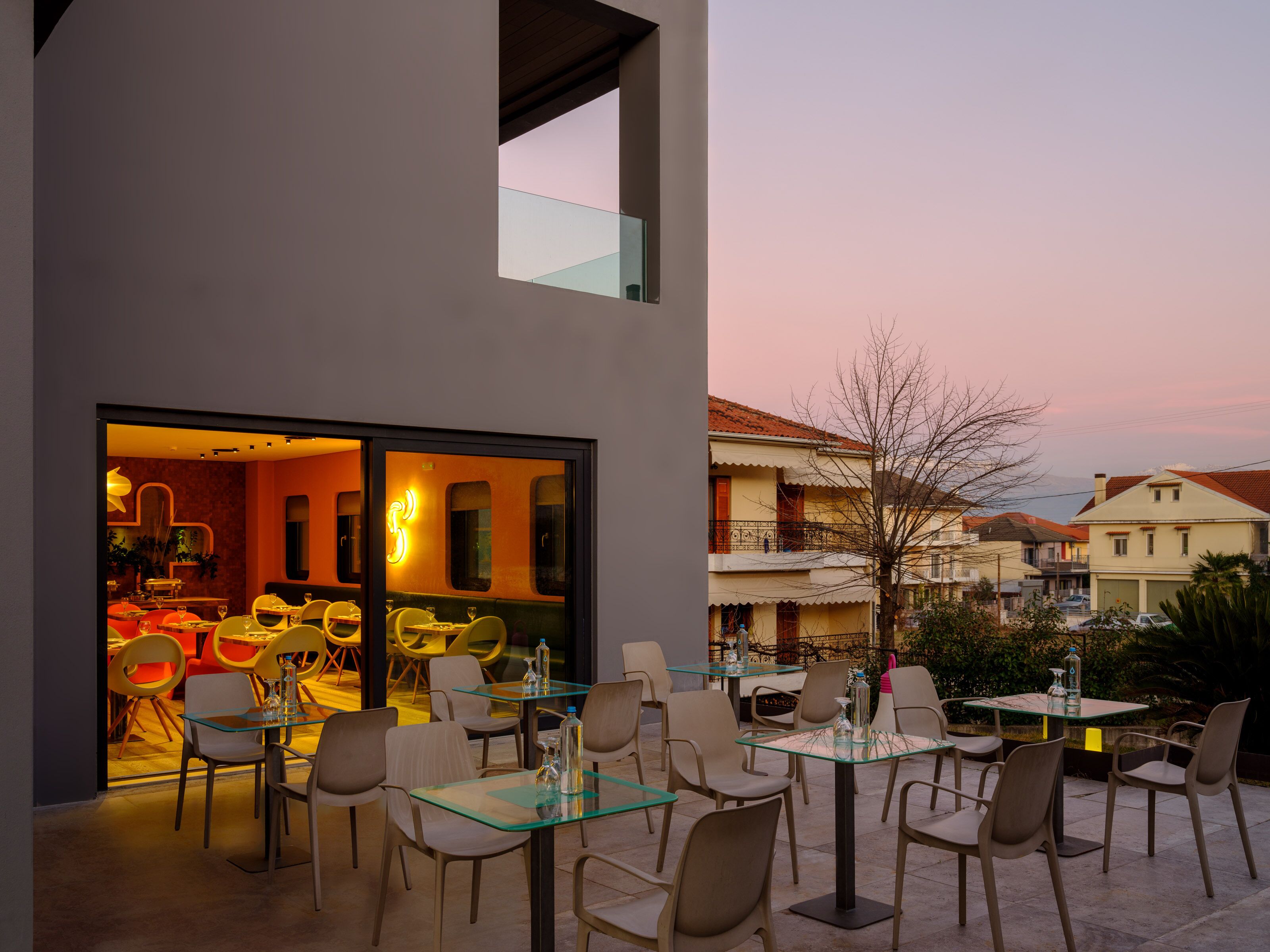 An outdoor dining area with glass tables and chairs, a view of a restaurant inside, and surrounding residential buildings.