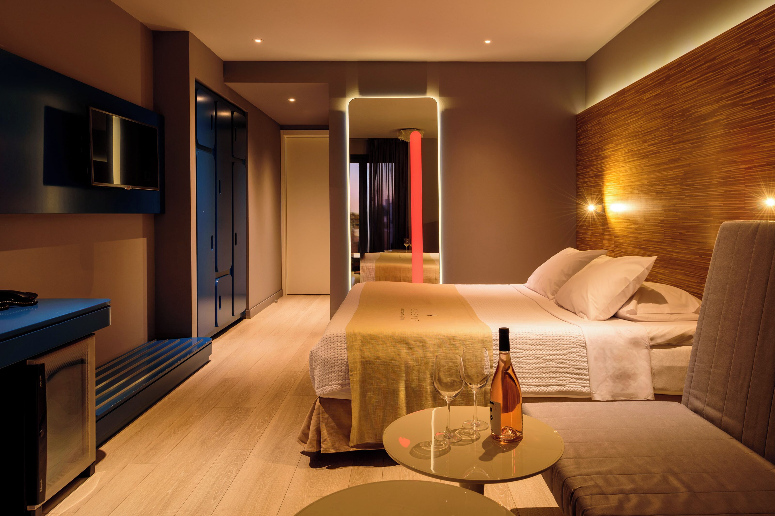 Ein modernes Hotelzimmer mit Bett, Fernseher, Minibar, Tisch mit Gläsern und einer Flasche.
