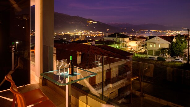 Un balcón con una mesa de vidrio, dos copas de vino y una botella. El balcón tiene vistas a un paisaje urbano con edificios y montañas al fondo.