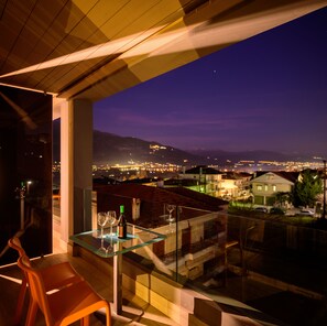 Un balcón con una mesa de vidrio, dos copas de vino y una botella. El balcón tiene vistas a un paisaje urbano con edificios y montañas al fondo.