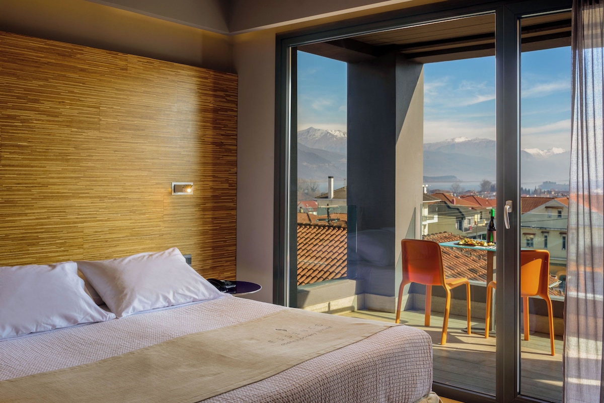 A modern hotel room with a large bed, a wooden accent wall, and a balcony with a view of the city and mountains.
