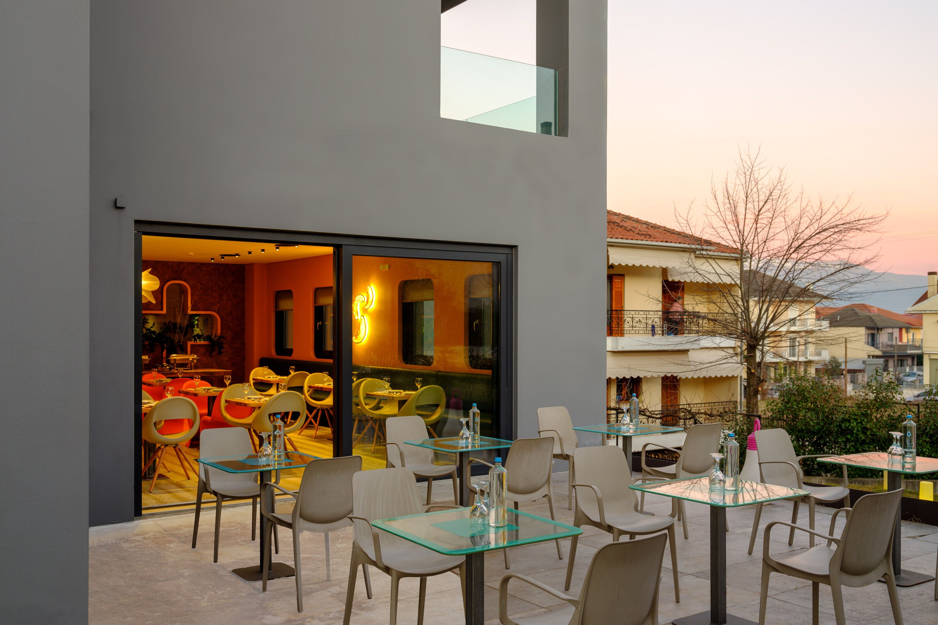 An outdoor dining area with glass tables and chairs, a view of the interior restaurant, and a building with balconies in the background.