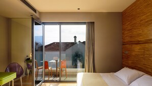 Una habitación de hotel moderna con una cama grande, una puerta de vidrio que da a un balcón, una mesa pequeña con sillas y vistas a la ciudad.