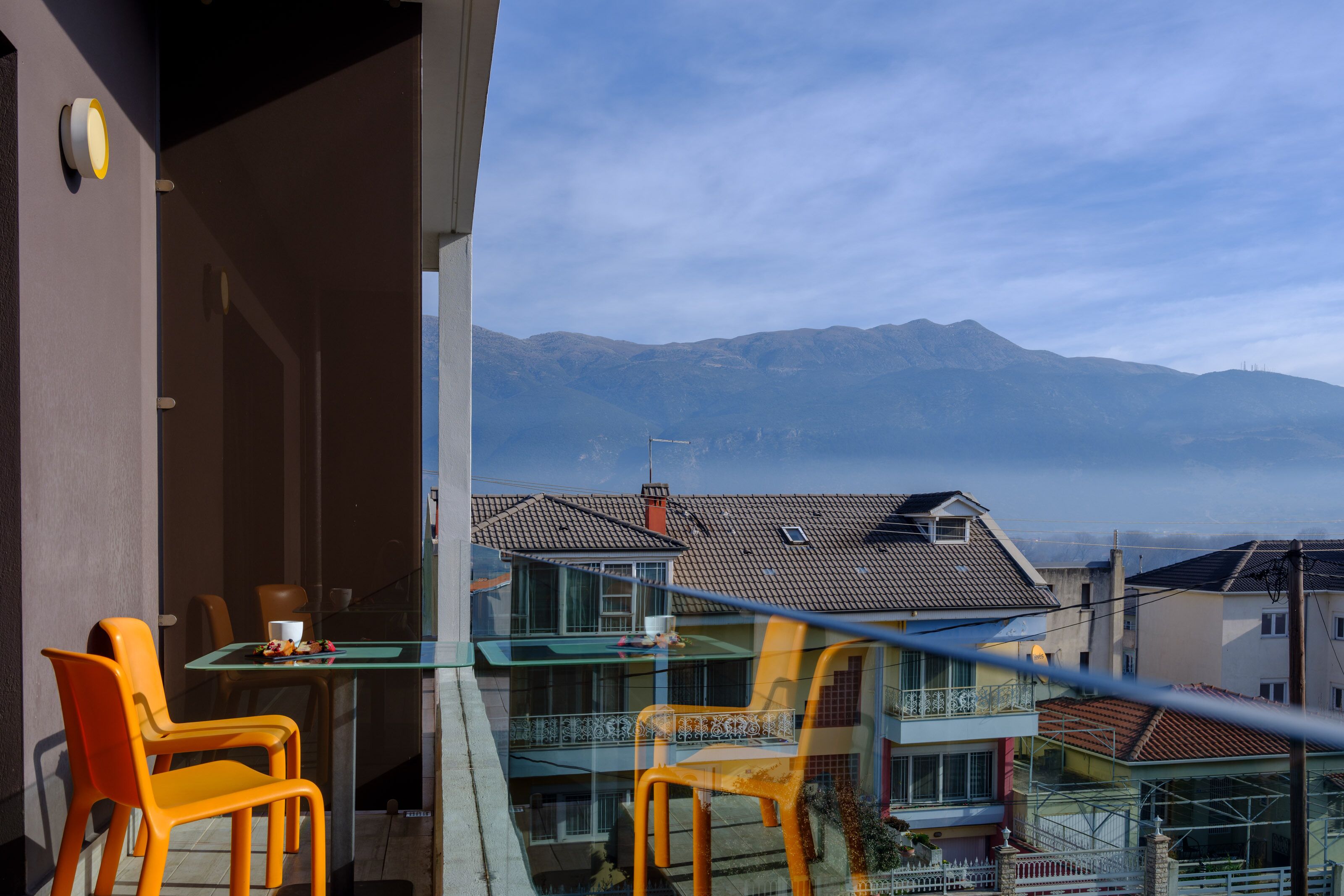 Un balcón con sillas naranja y una mesa, con vistas a una zona residencial con casas y montañas al fondo.