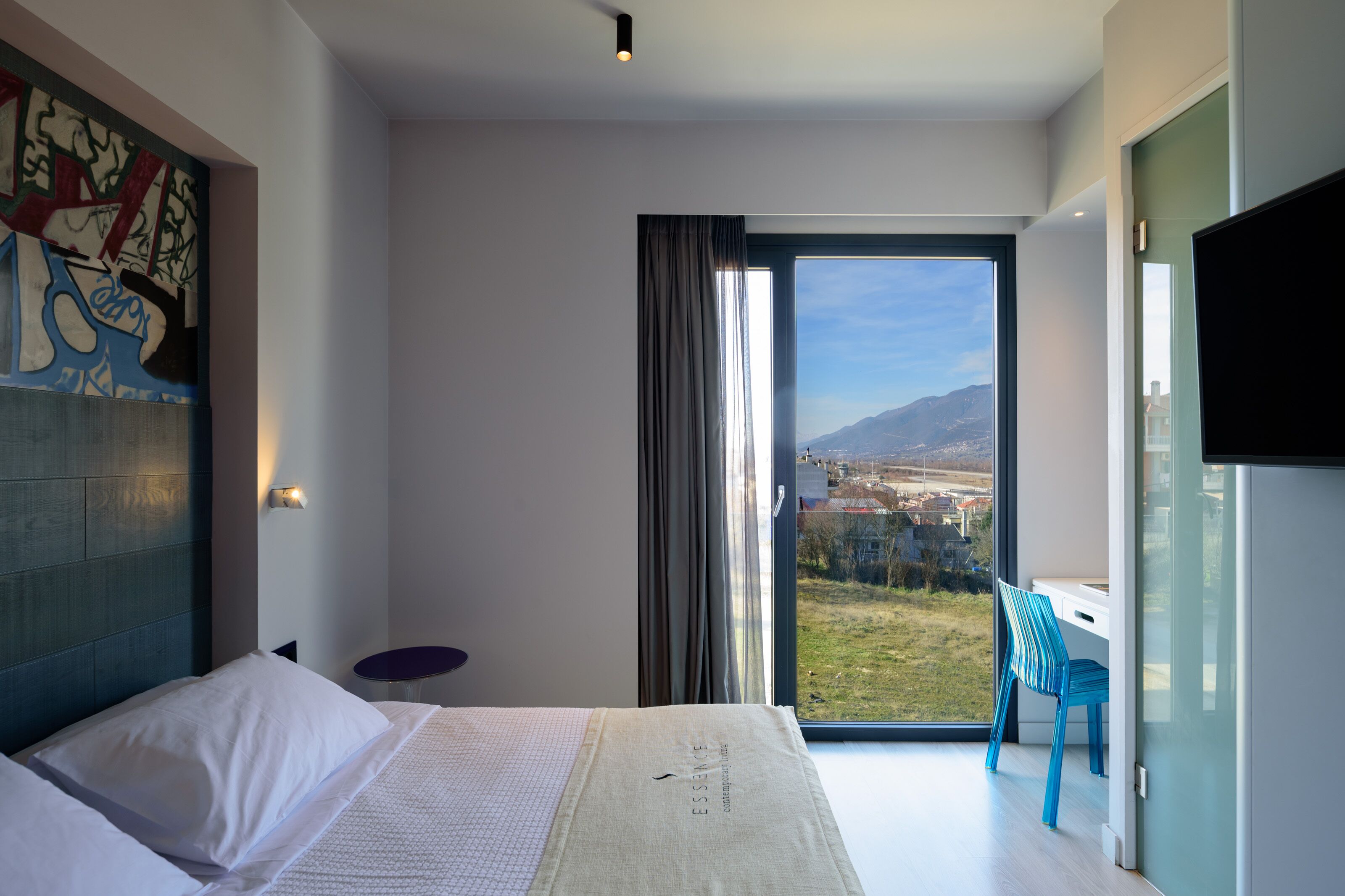 Una camera d'albergo moderna con un letto grande, una scrivania e una sedia vicino a una porta scorrevole in vetro che offre vista su un paesaggio urbano e montagne.