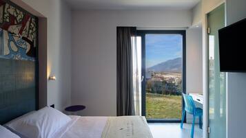 Una camera d'albergo moderna con un letto grande, una scrivania e una sedia vicino a una porta scorrevole in vetro che offre vista su un paesaggio urbano e montagne.