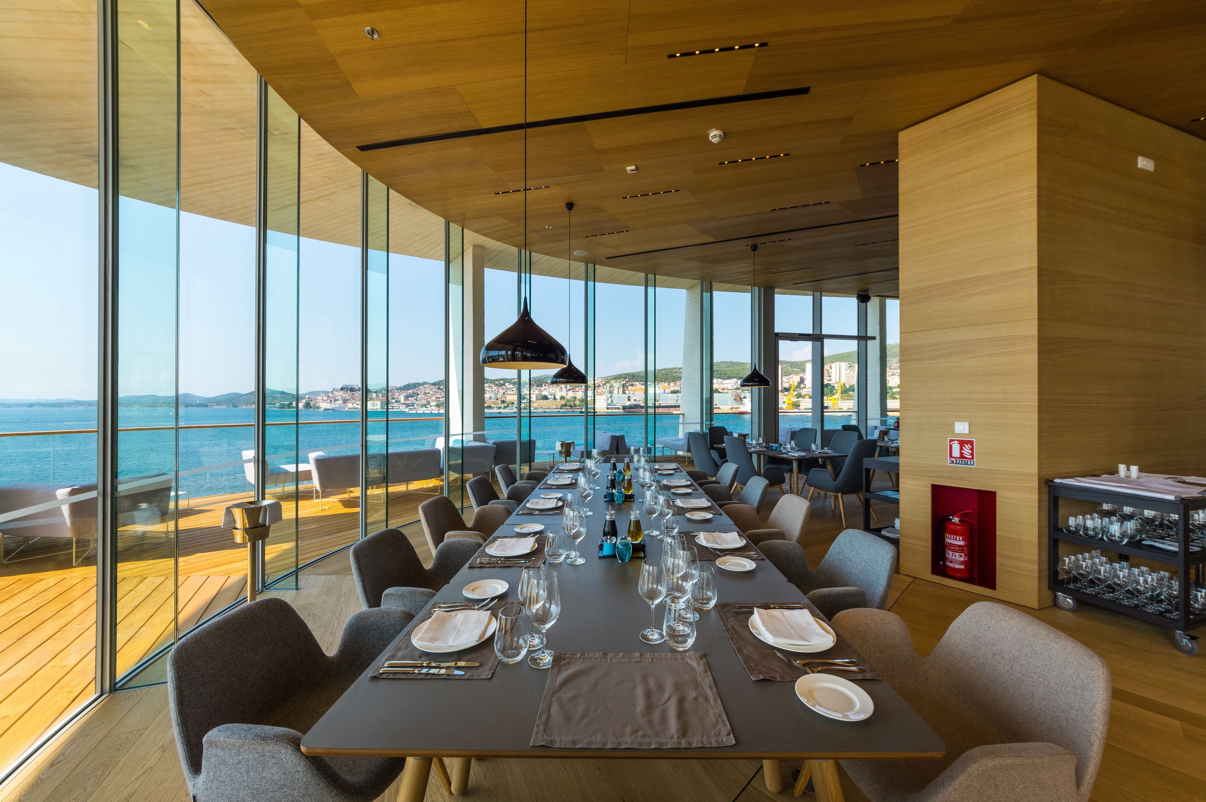 A dining area with a long table set for a meal, large windows offering a view of the sea, and a fire extinguisher on the wall.