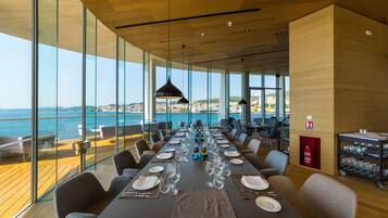 Un comedor con una larga mesa dispuesta para una comida, grandes ventanales con vista al mar y un extintor de incendios en la pared.