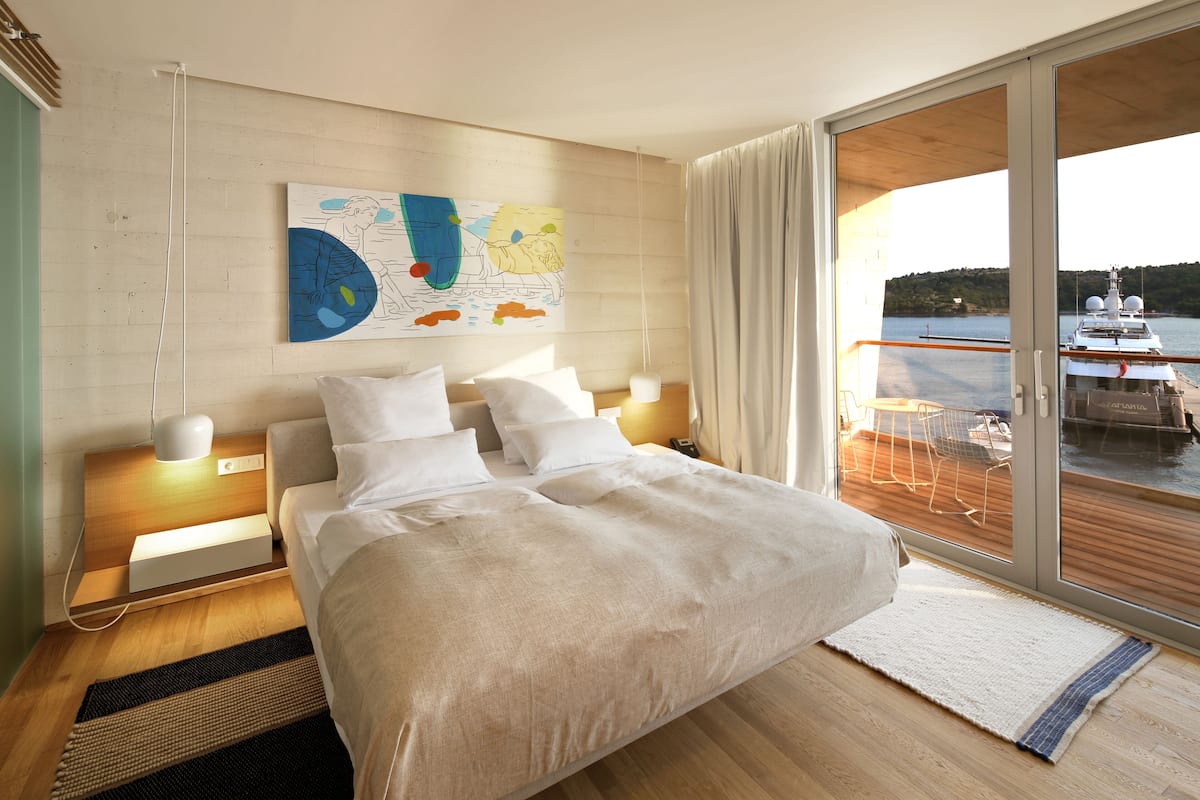 A modern hotel room with a large bed, a wooden headboard, a map artwork on the wall, and a view of a boat through a sliding glass door.