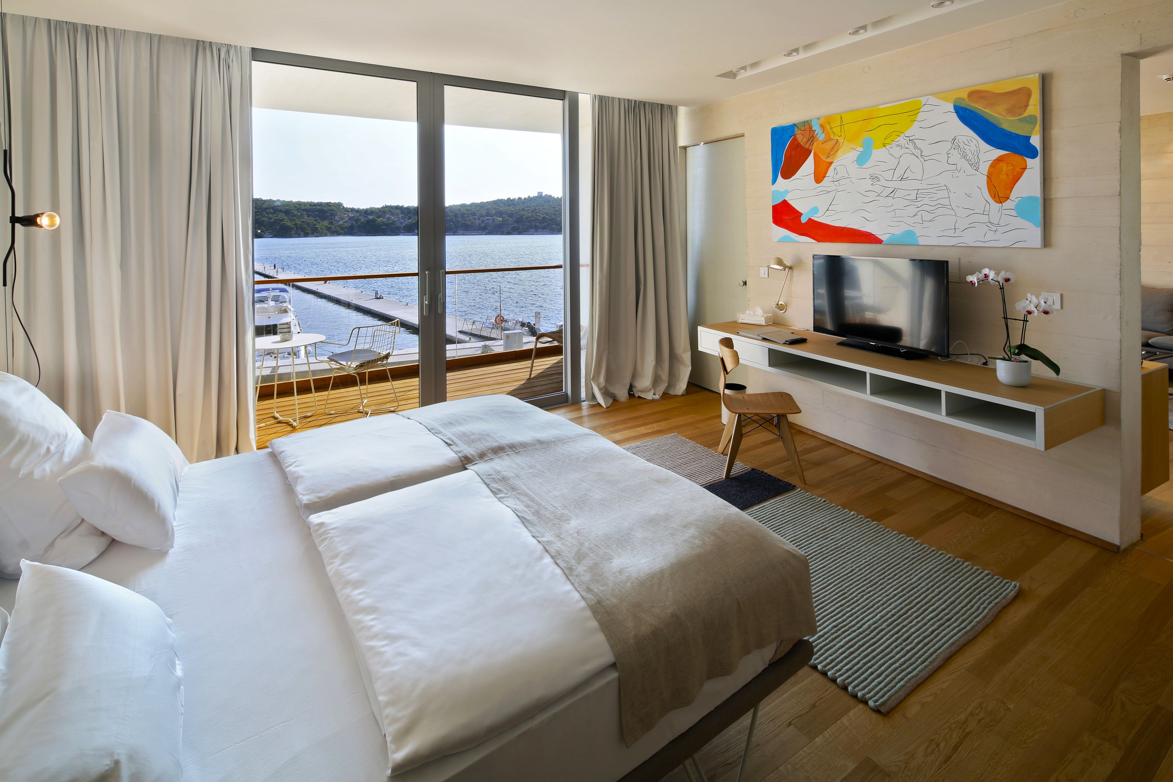 A modern hotel room with a large bed, a TV, a desk, and a view of a lake.