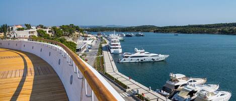 A waterfront promenade with a wooden deck, boats docked along the shore, and a clear view of the water and distant hills.