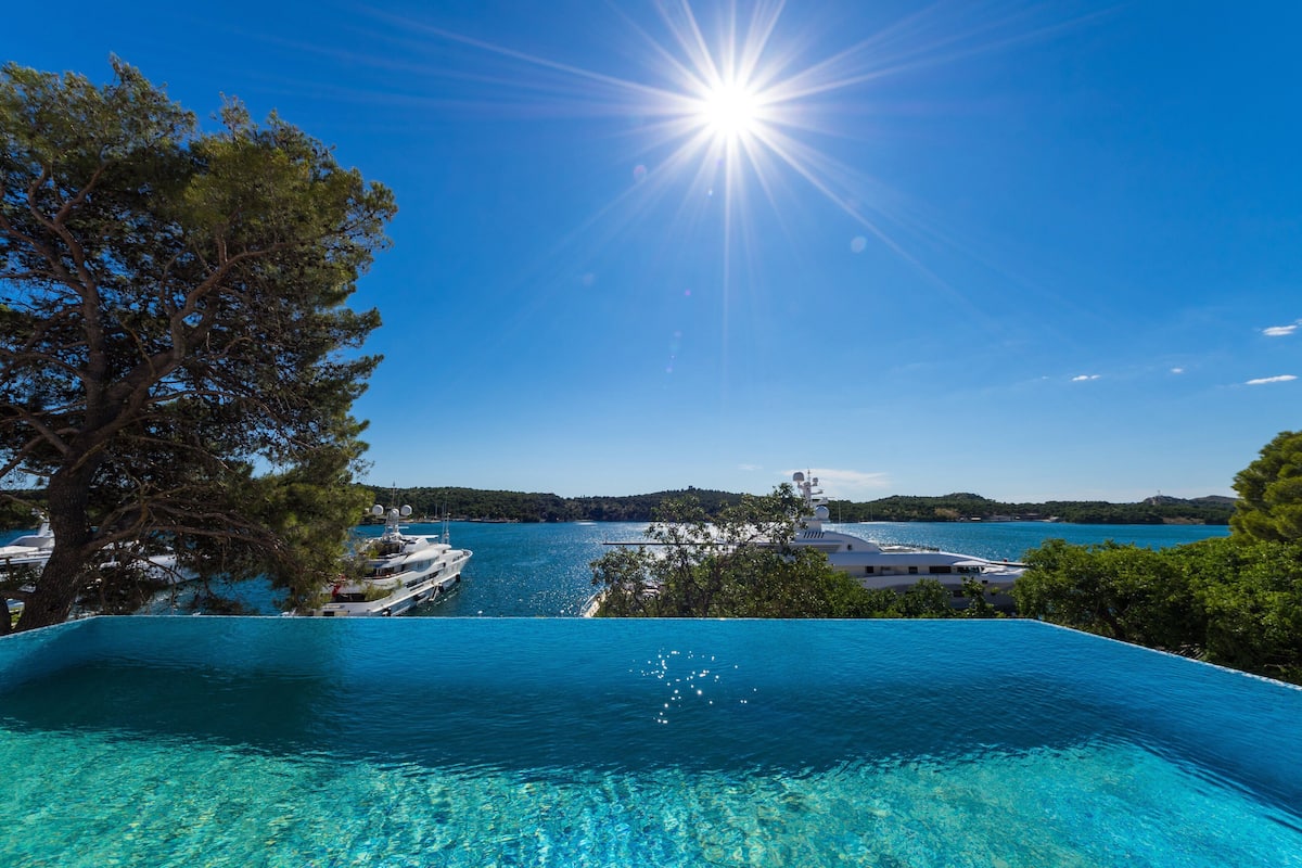 An infinity pool with clear blue water, a boat docked nearby, and a view of a coastal landscape with trees and hills.