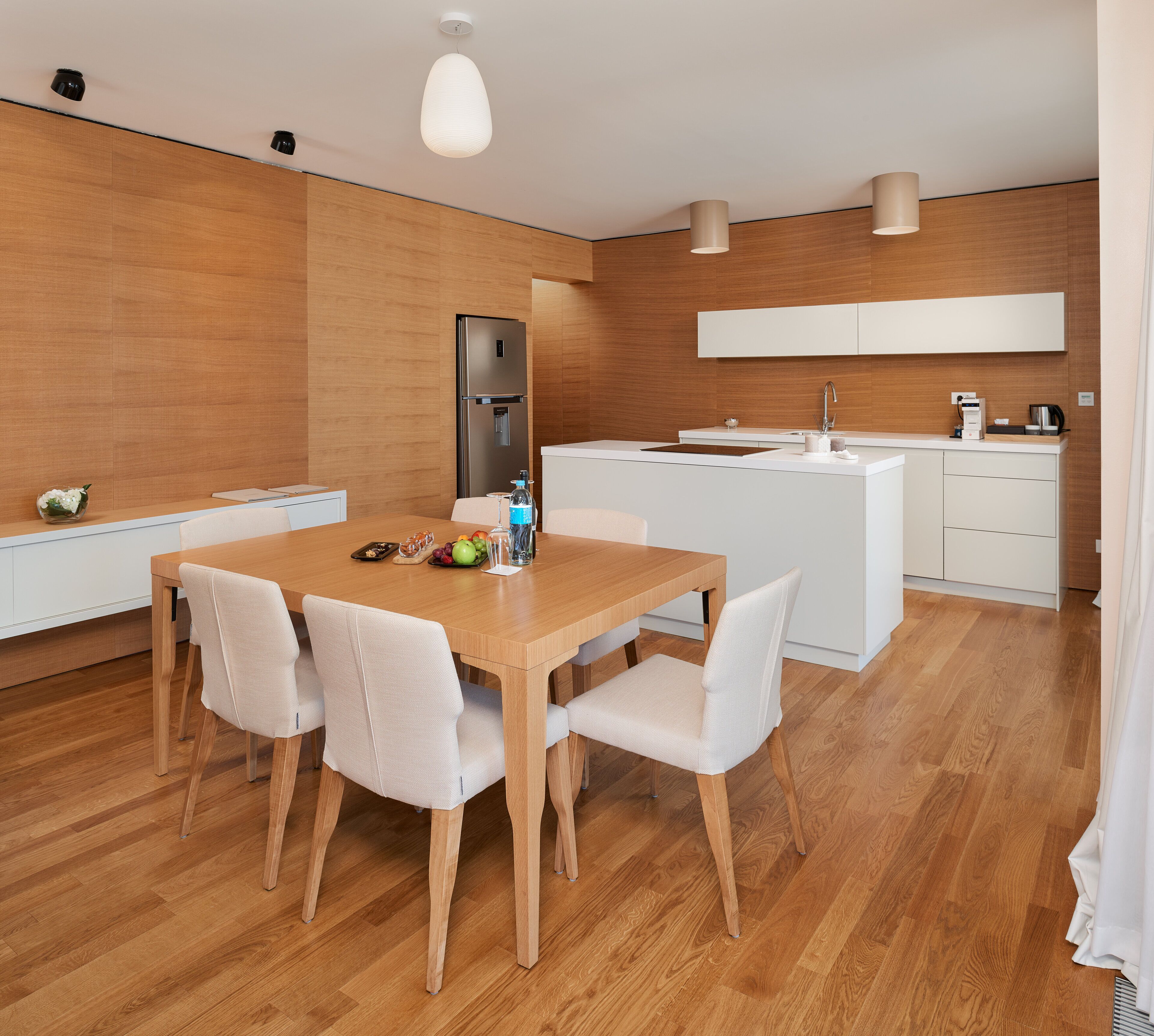 A modern kitchen with a wooden dining table, white chairs, and a built-in refrigerator.