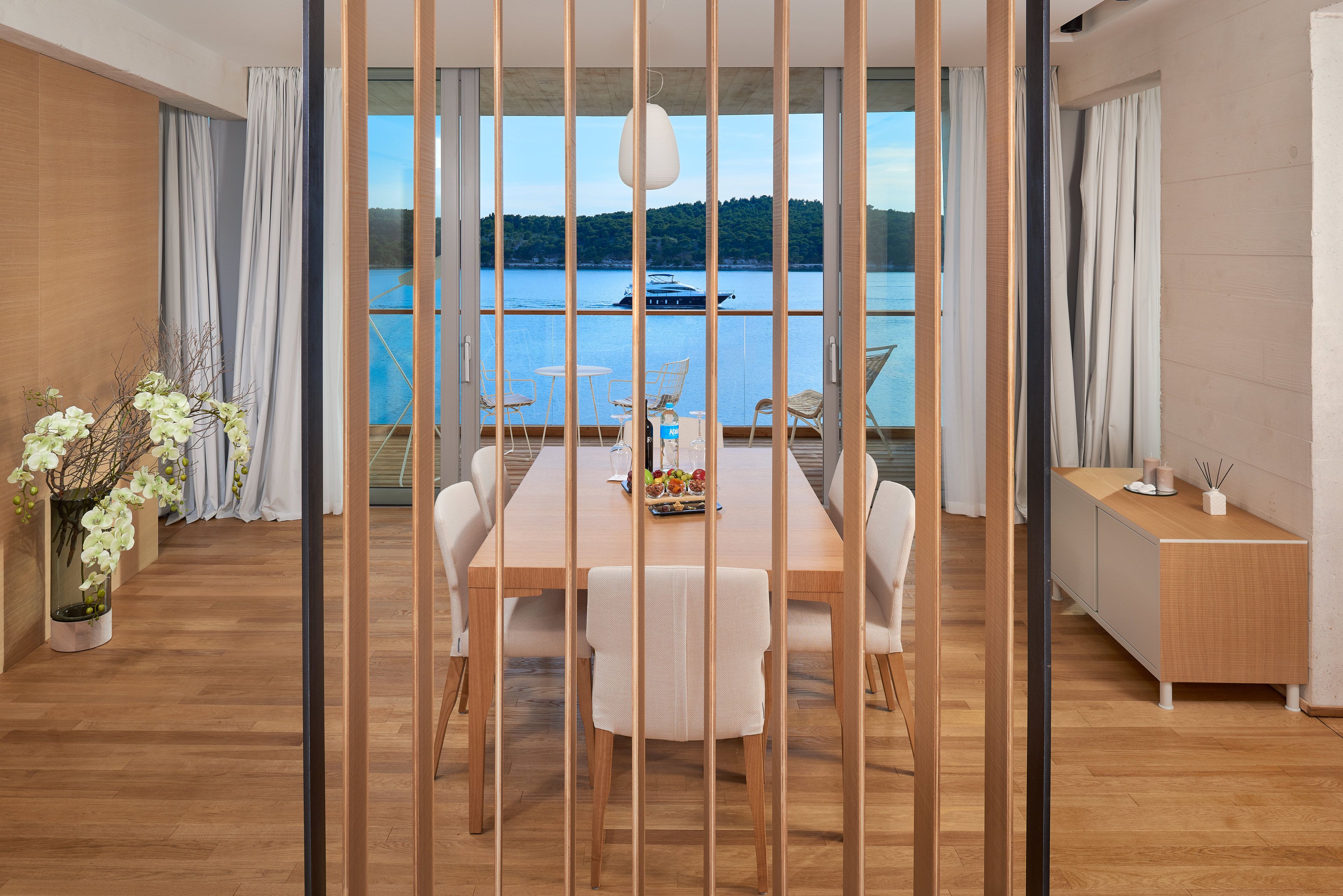 Un comedor moderno con vistas a un lago y un bote, suelo de madera, sillas blancas y una mesa de madera con un arreglo de frutas.