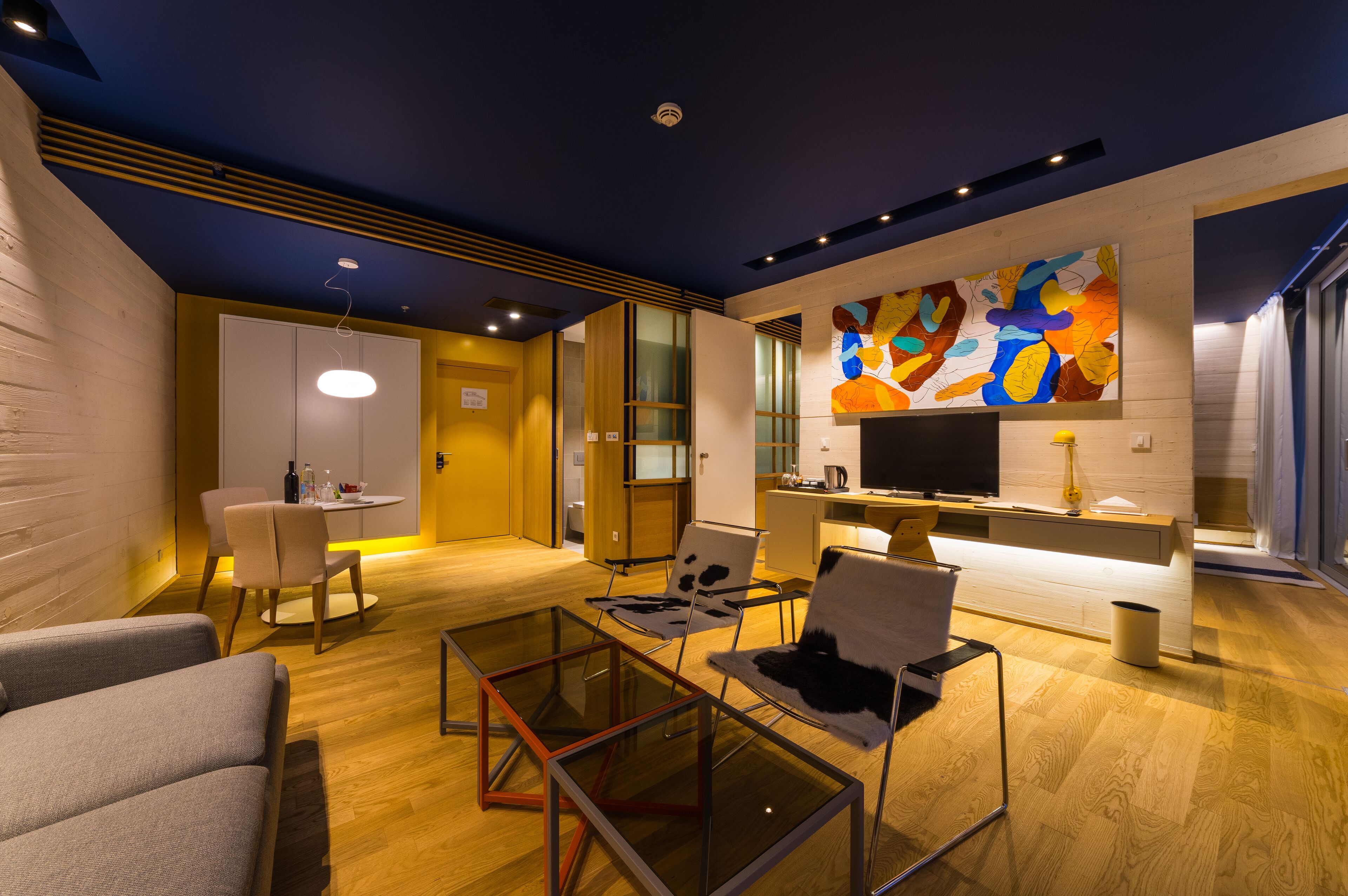 Ein modernes Wohnzimmer mit einem Glastisch, zwei Sofas, einem Fernseher und einem farbenfrohen, abstrakten Gemälde an der Wand.