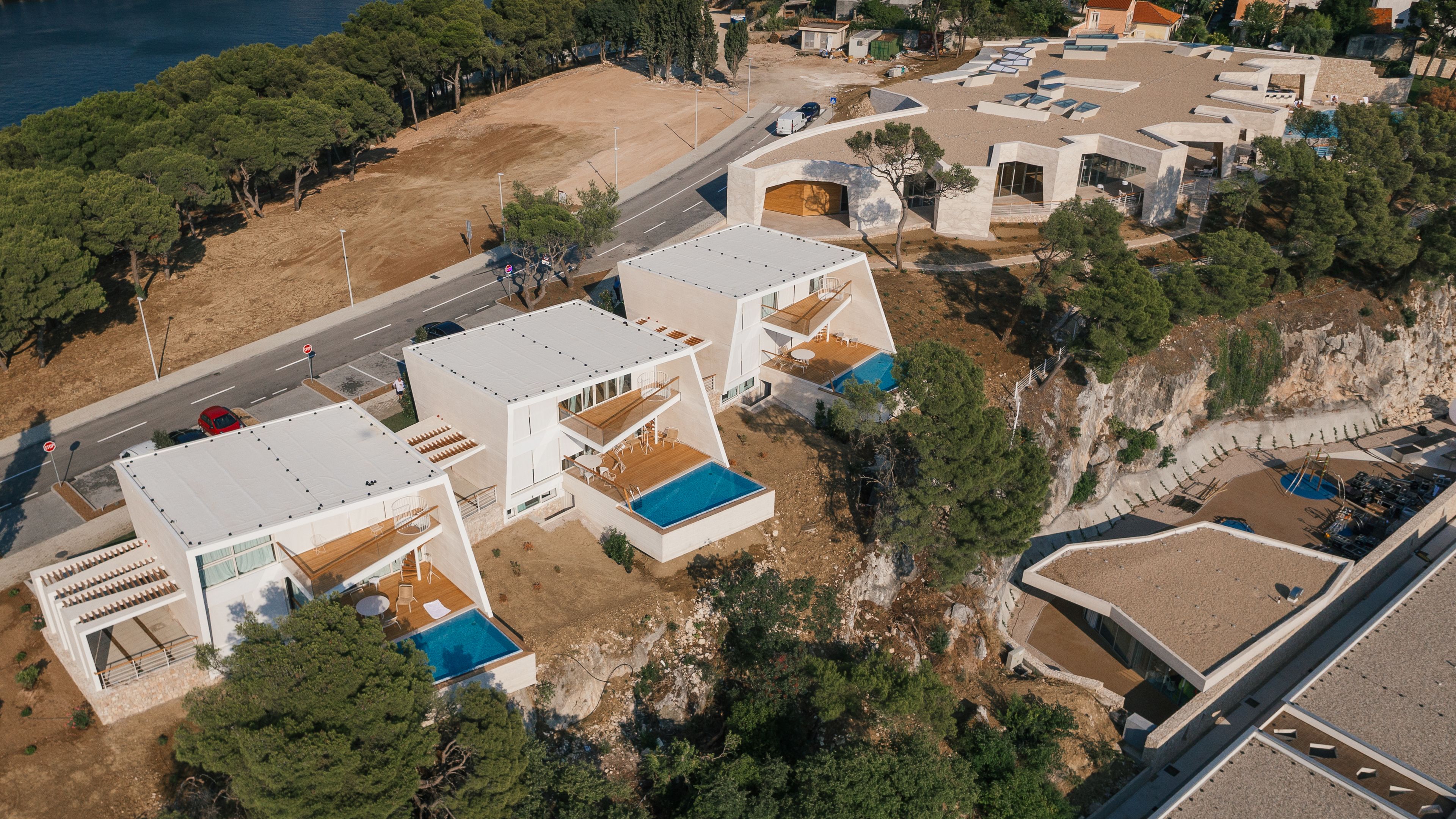 Vue aérienne d’une propriété située sur la côte, dotée d’une architecture moderne, comprenant plusieurs bâtiments à toits plats, une piscine et une route à proximité.
