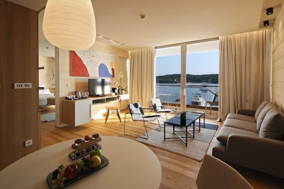 A modern living room with a sofa, a dining table with fruit, a TV, and a view of a lake.