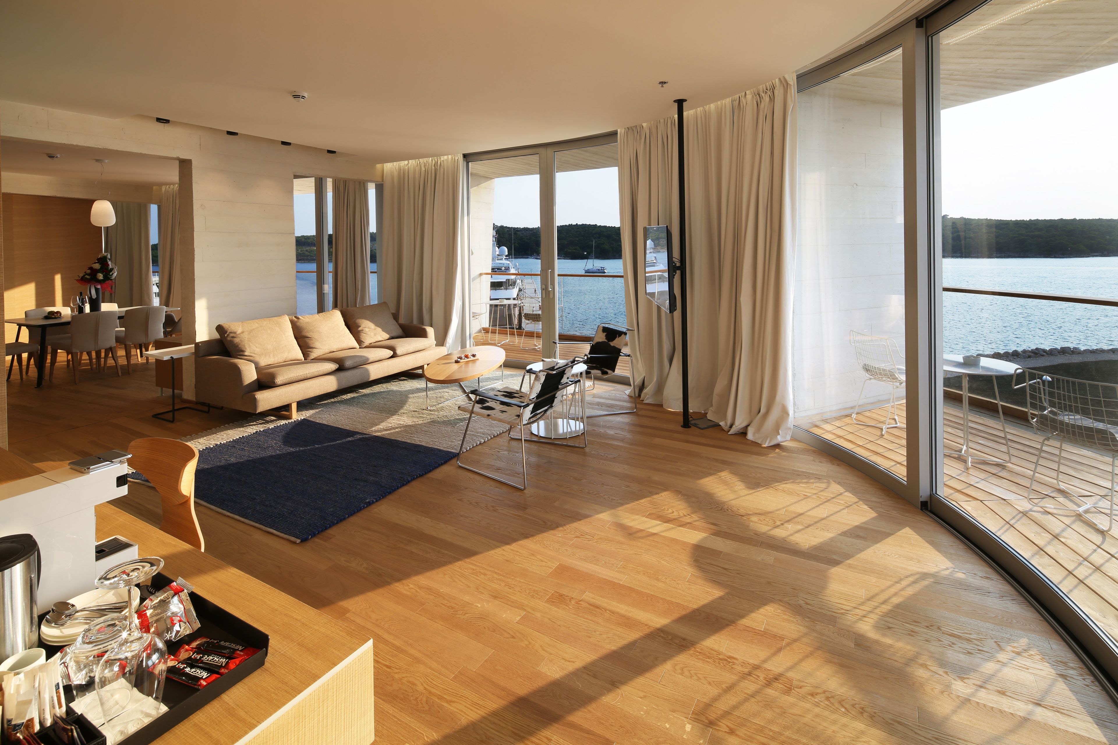 A spacious living room with a large sofa, a dining area, and a balcony with a view of the water.