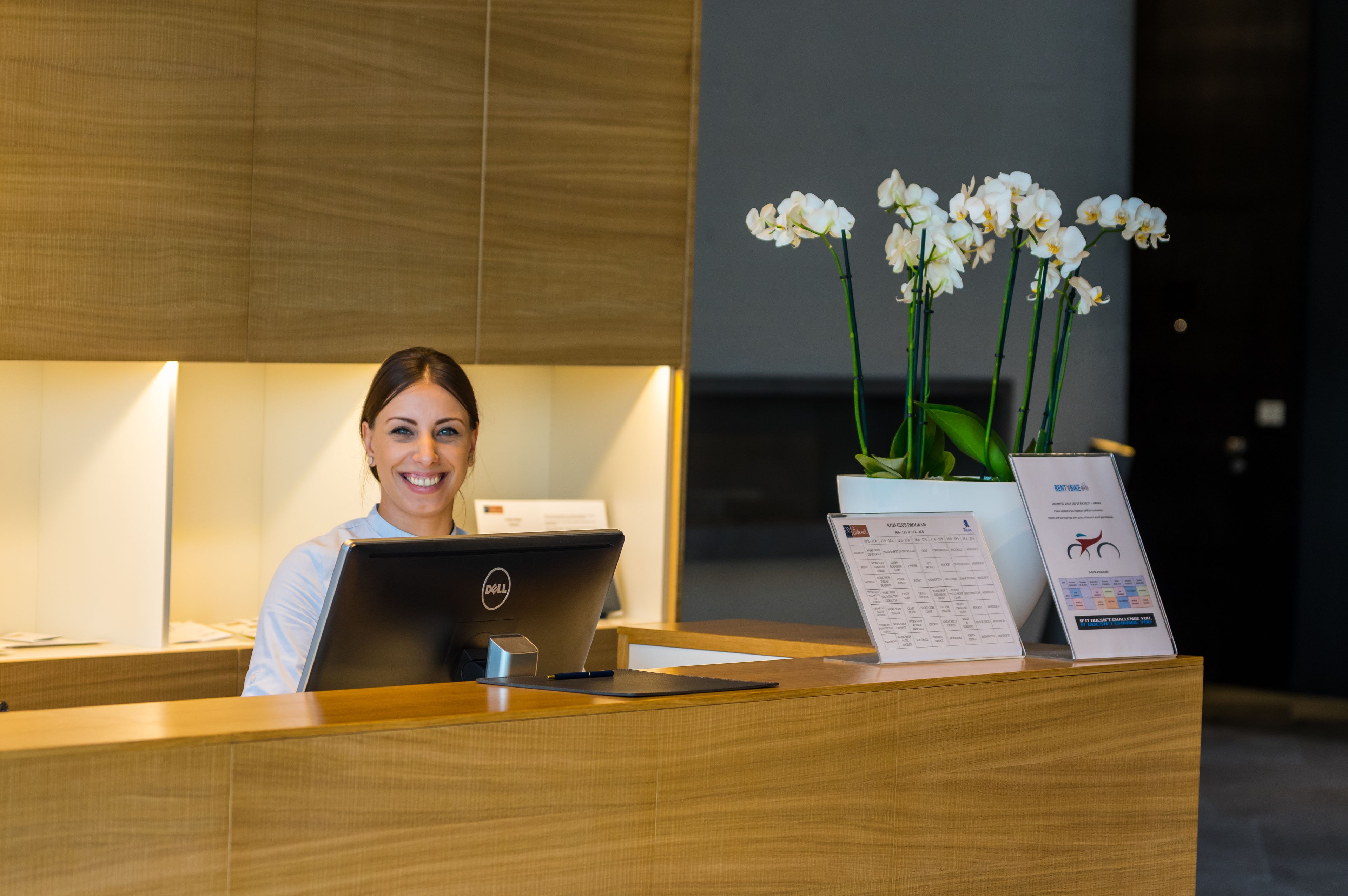 Una mesa de recepción con una computadora portátil Dell, una orquídea en maceta y un cartel de bienvenida.