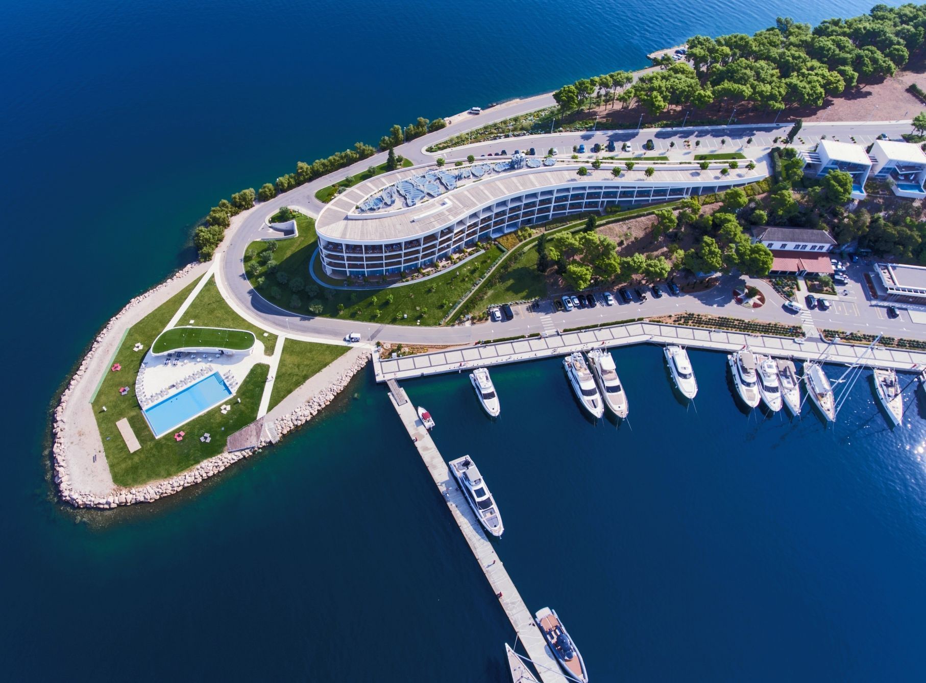 Eine Luftaufnahme eines Anwesens am Wasser mit einem Hafen, einem Schwimmbad und einem Gebäude mit kurvem Verlauf und modernem Design.