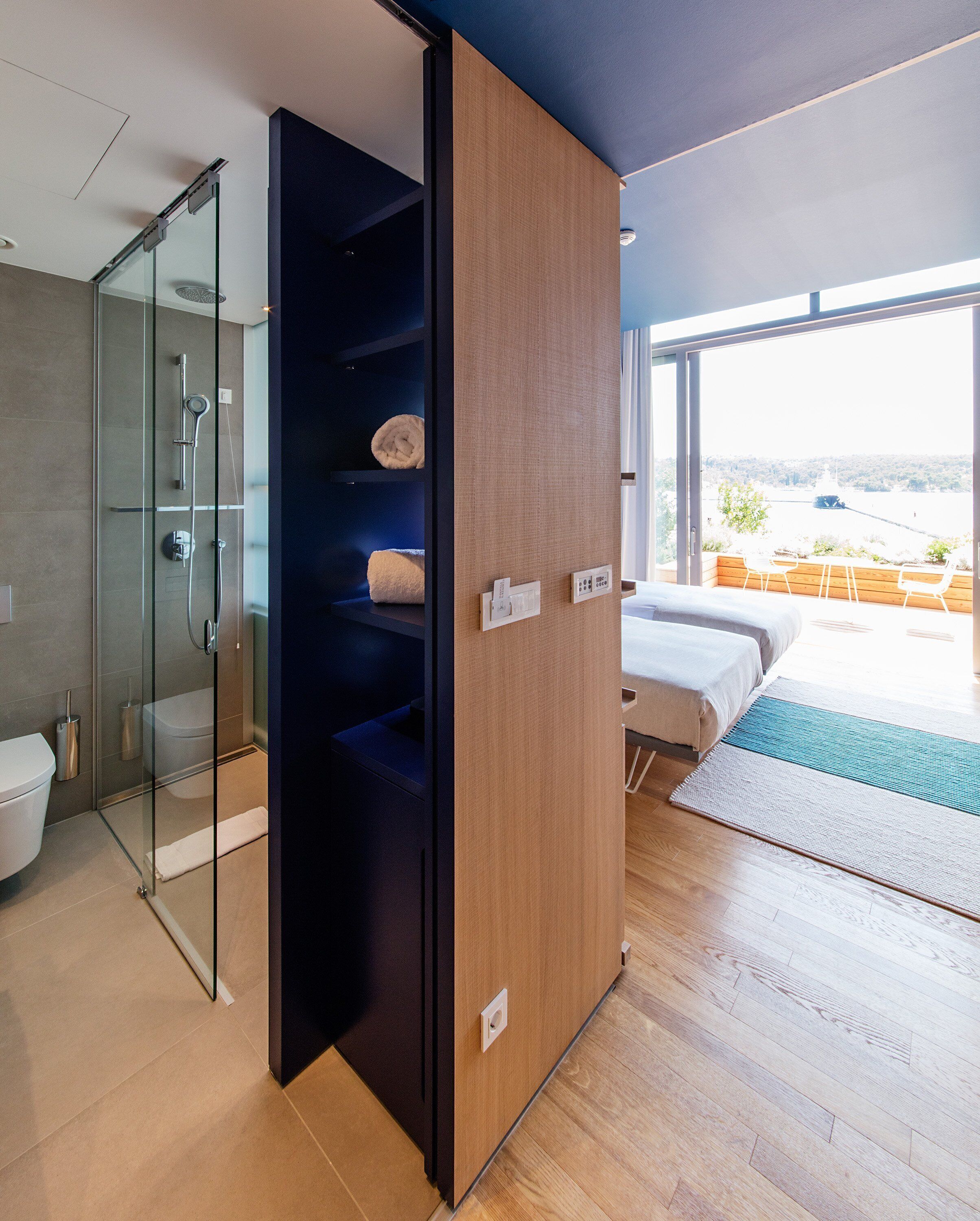 A modern hotel room with a glass shower enclosure, a wooden wardrobe, a bed with a view, and a small balcony with outdoor seating.