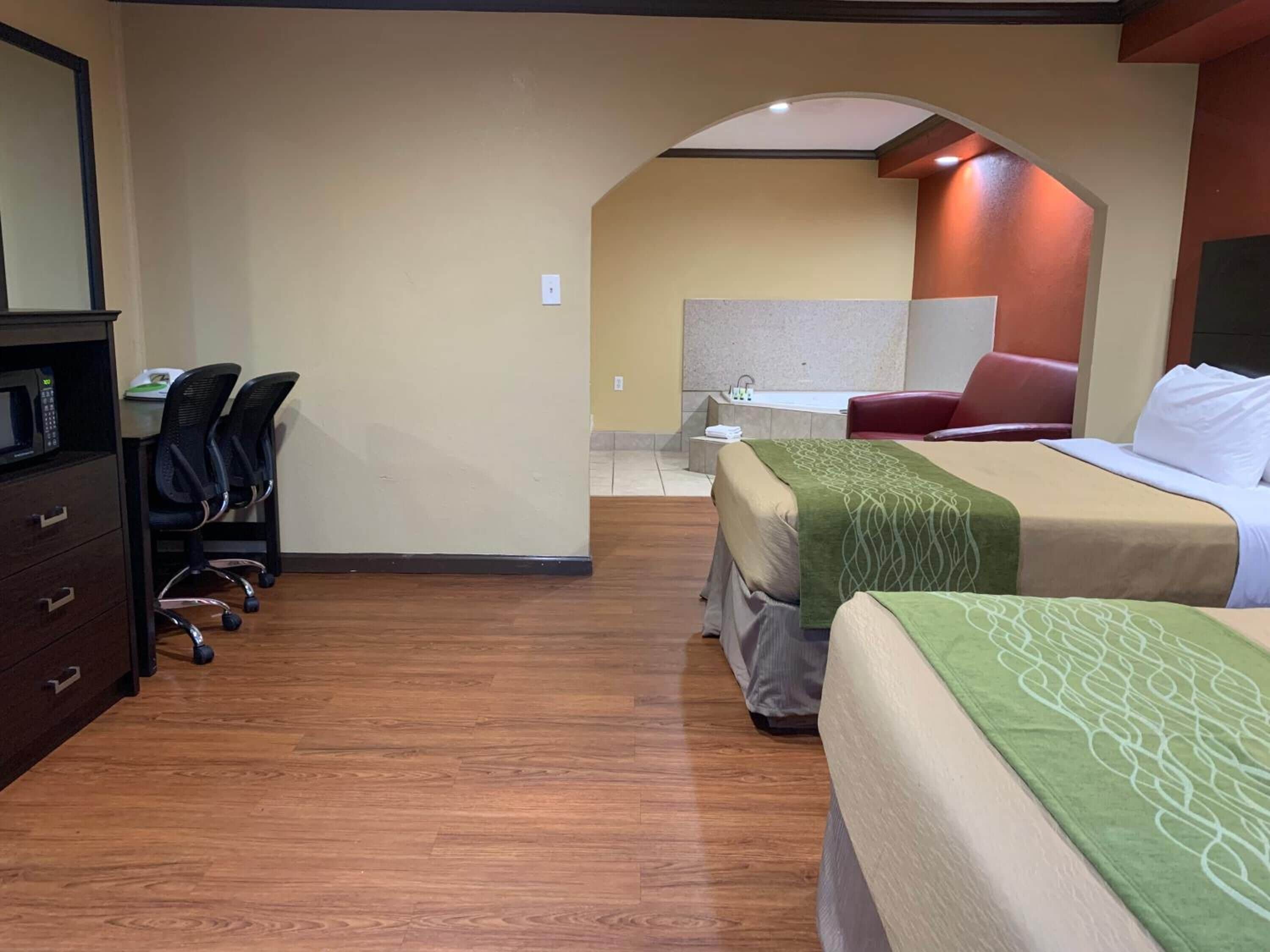 A hotel room with two beds, a desk with chairs, a microwave, and a bathroom visible through an archway.
