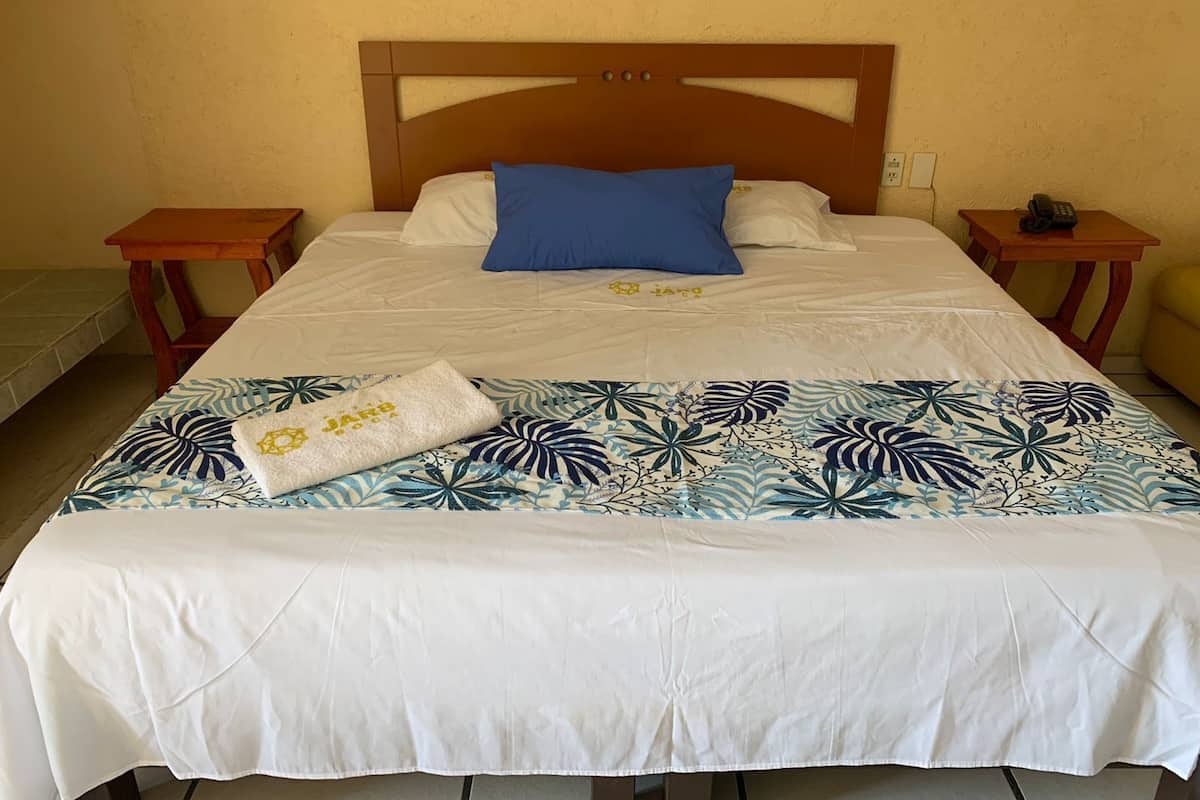 Ein ordentlich bezogenes Bett mit einer blumigen Tagesdecke, einem blauen Kopfkissen und einem weißen Handtuch mit Aufdruck. Es gibt zwei Nachttische, auf einem davon steht ein Telefon.