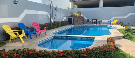 Piscina con sillones de descanso cercanos y una pequeña mesa, rodeada de follaje y flores.