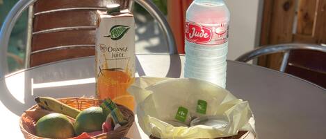 A table with a juice carton, a bottle of water, a basket of fruits, a packet of cookies, and a loaf of bread.