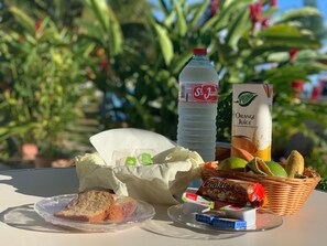 A table with a plate of food, a bottle of water, a carton of orange juice, a basket of fruits, and a package of cookies. - Hôtel Douce Vague (Sainte-Luce)
