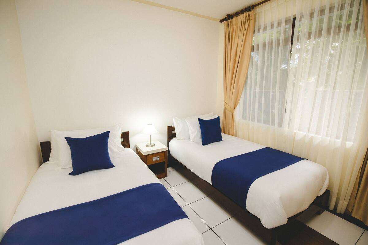 Een hotelkamer met twee enkele bedden, elk voorzien van een blauwe sprei en witte kussens. Tussen de bedden staat een nachtkastje met een lamp, en er is een raam met dunne gordijnen.