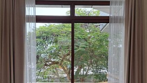 Ein Zimmer mit großem Fenster, durchscheinenden Vorhängen, Blick auf einen Baum und einem Balkongeländer.