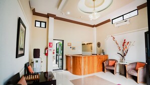 Eine Hotelhalle mit einer Rezeption, zwei Sesseln, einem Schachbrett, einem Feuerlöscher und einer Tür zum Außenbereich.