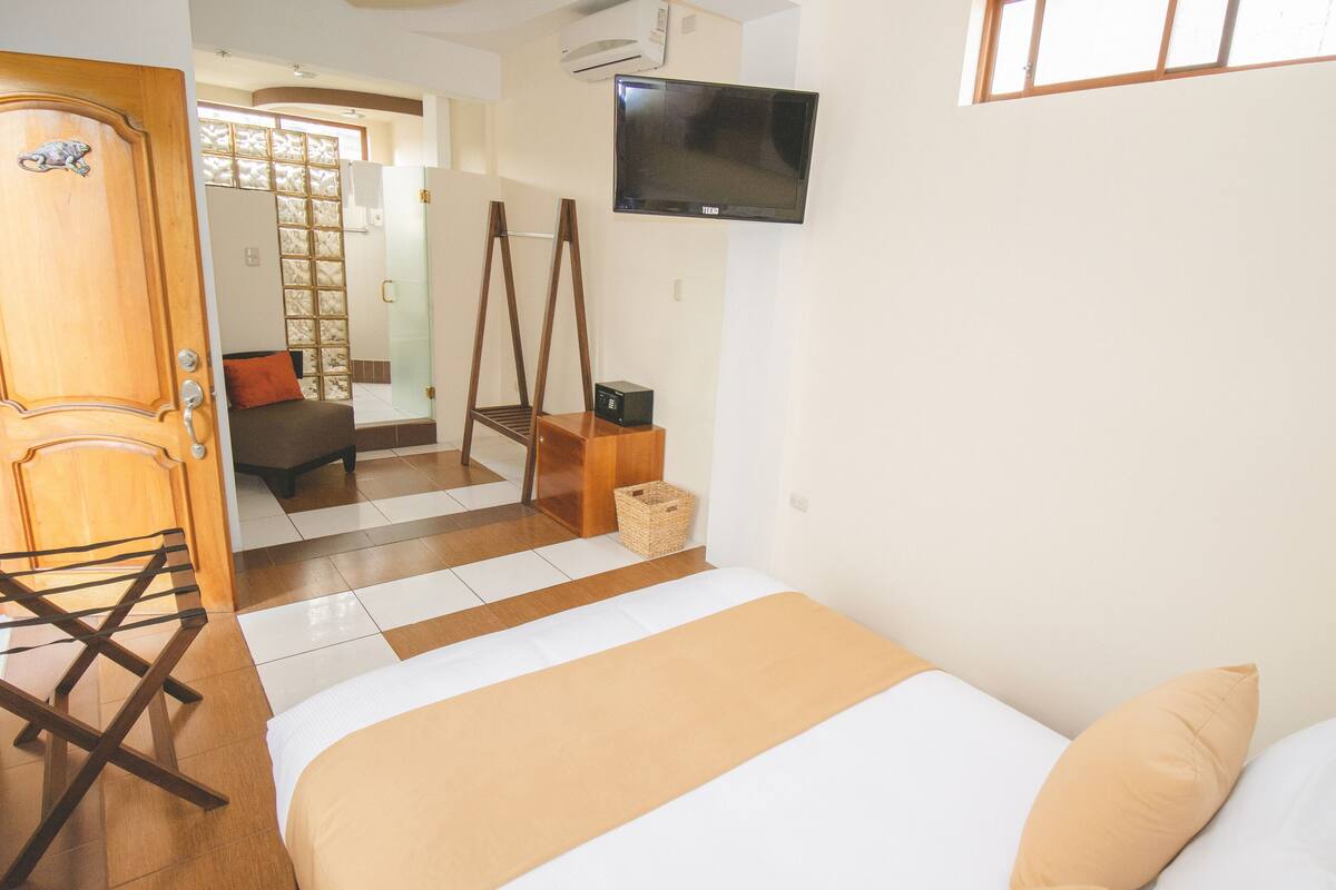 Een hotelkamer met een bed, een aan de muur bevestigde televisie, een houten stoel en een klein tafeltje met een mandje.