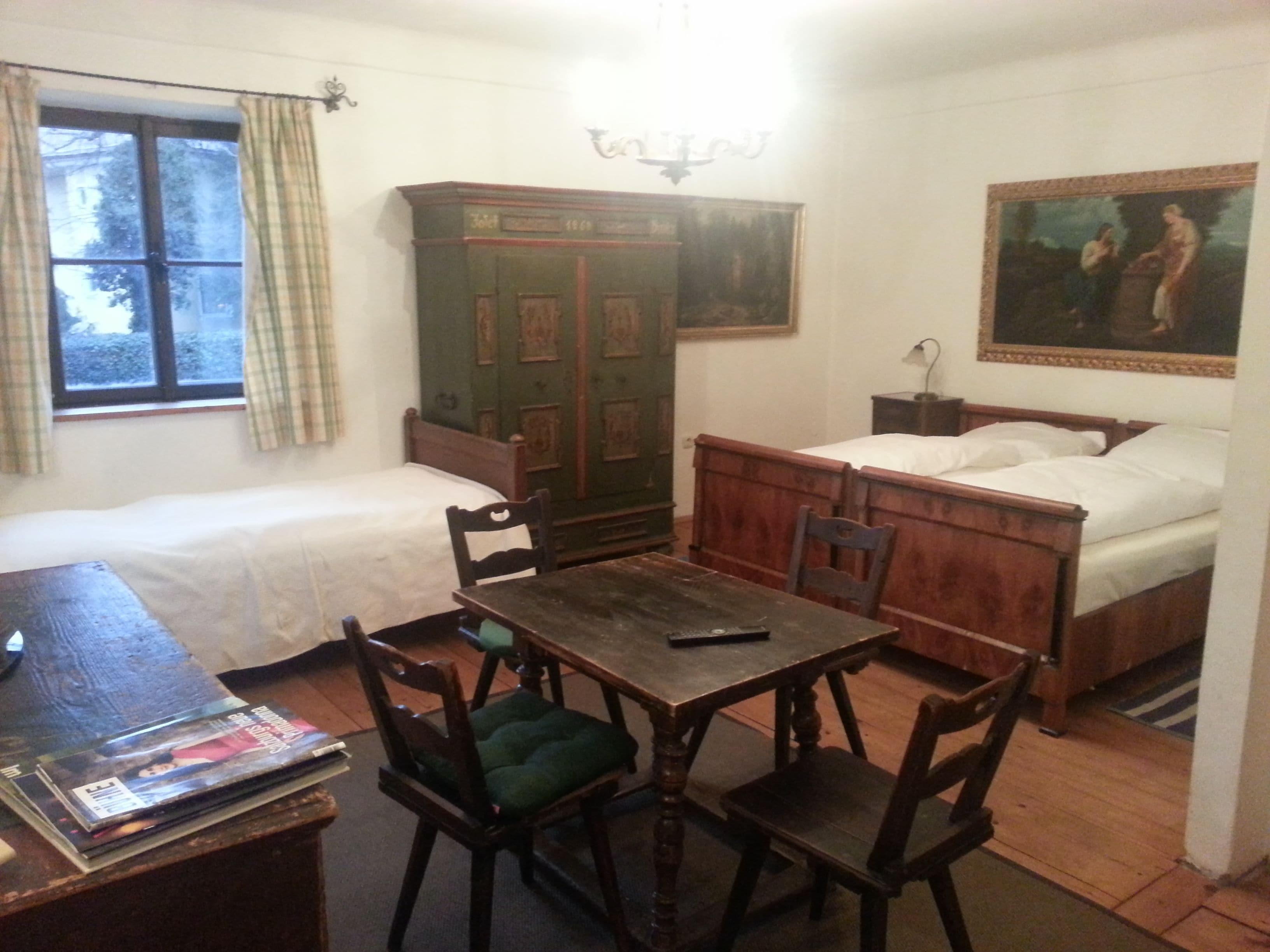 A room with two single beds, a wooden table with chairs, a desk, a green cabinet, and a painting on the wall.