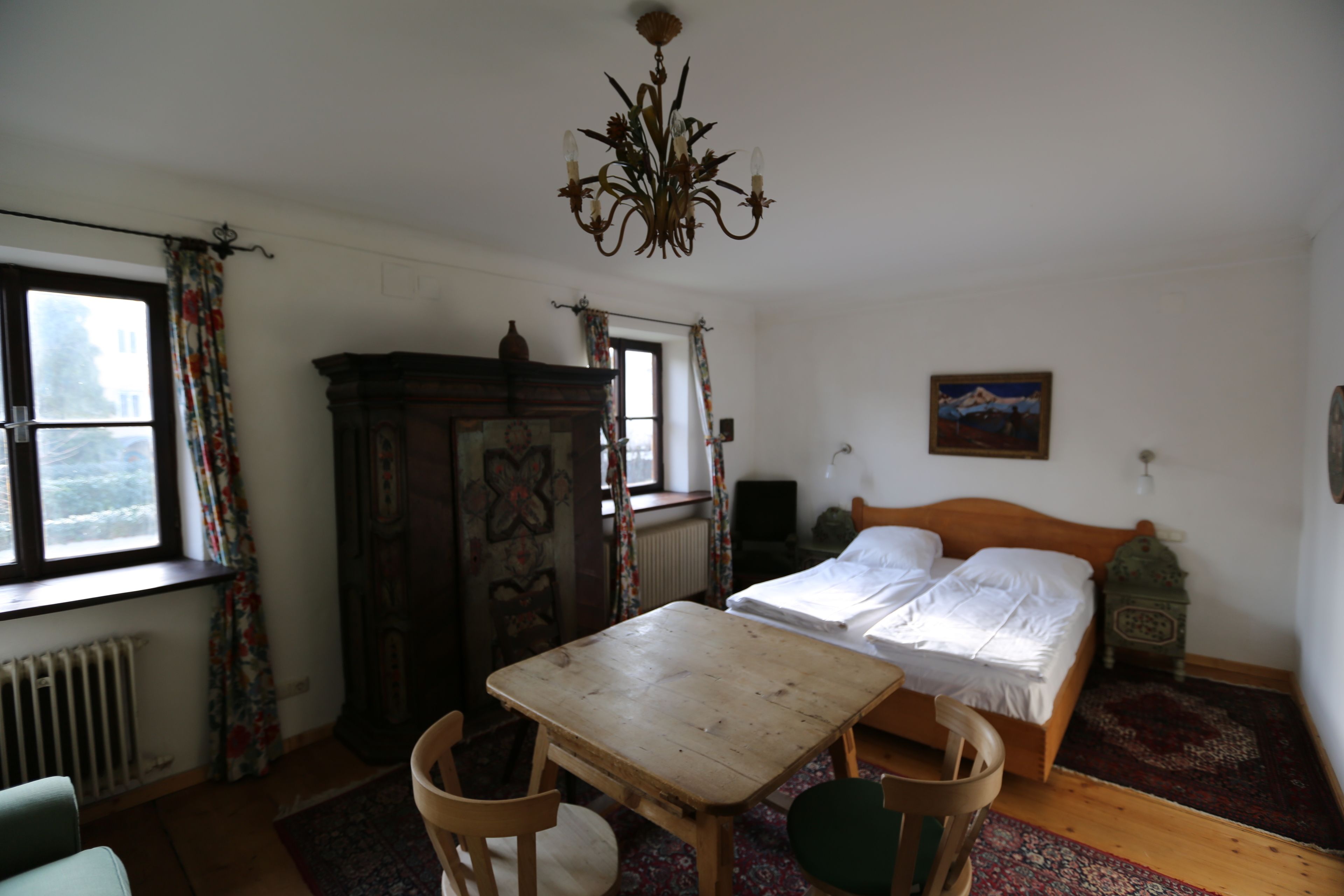 Een slaapkamer met een houten bed, een houten tafel, een stoel, een radiator, een raam met gordijnen en een kroonluchter.