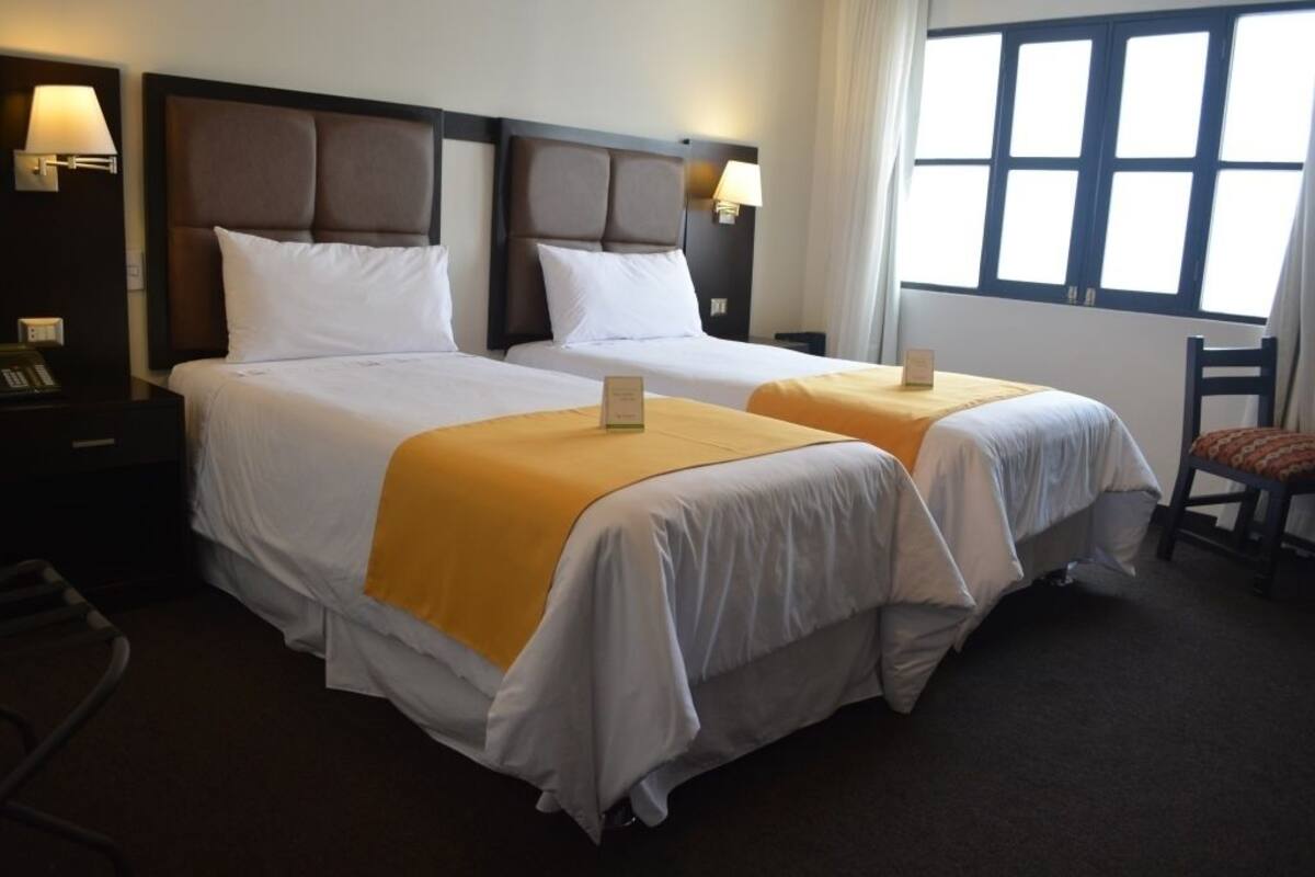 Ein Hotelzimmer mit zwei Betten, jeweils mit Kopfteil, weißer Bettwäsche und einem gelben Akzent. Es gibt Nachttischlampen, einen Sessel und ein Fenster mit Vorhängen.