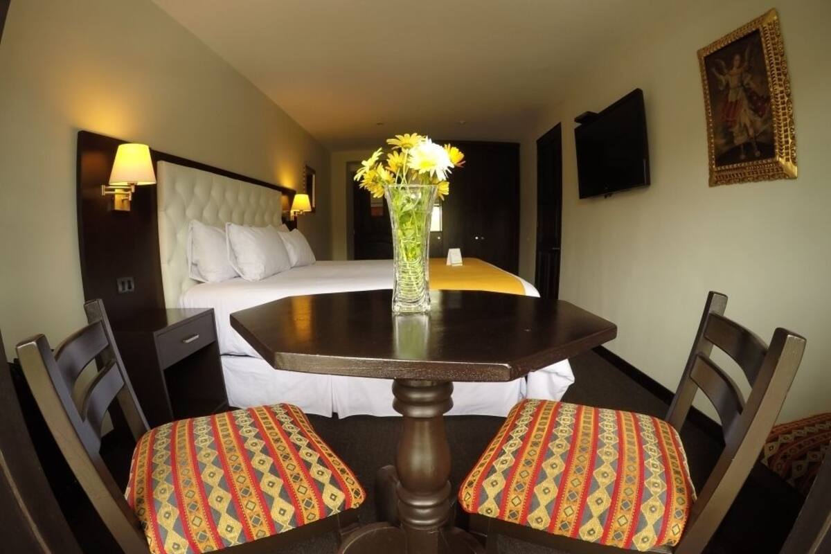 Ein Hotelzimmer mit Bett, Esstisch mit Stühlen, einer Blumenvase und einem Bild an der Wand.