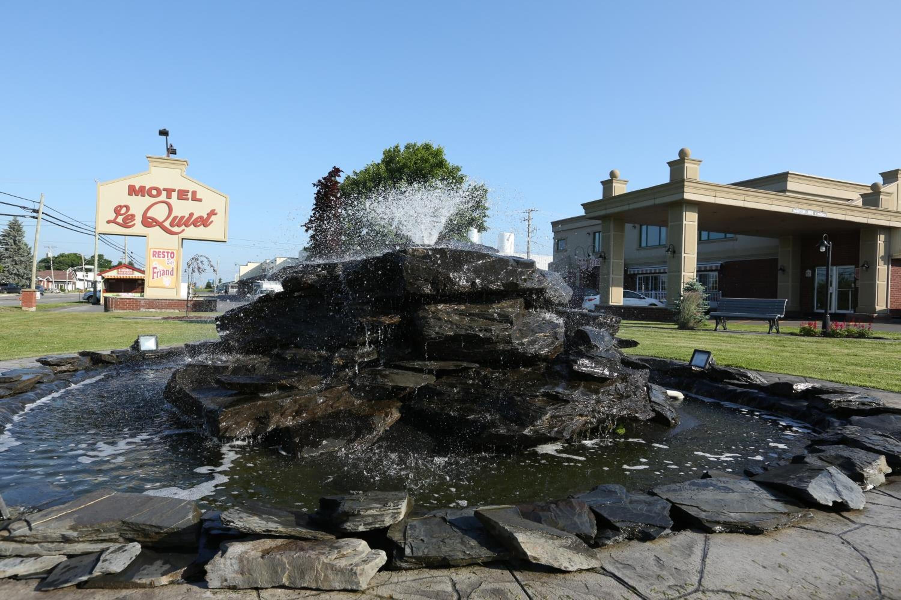 A fountain with water jets in a stone basin, surrounded by a paved area with a motel sign in the background.