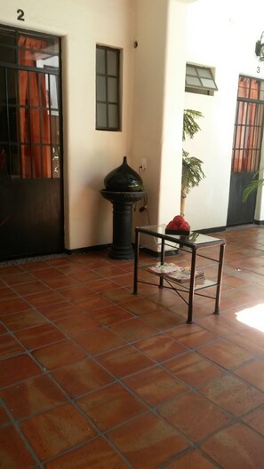 Un sol carrelé, une porte avec un numéro, une petite table avec un vase et une plante en pot.