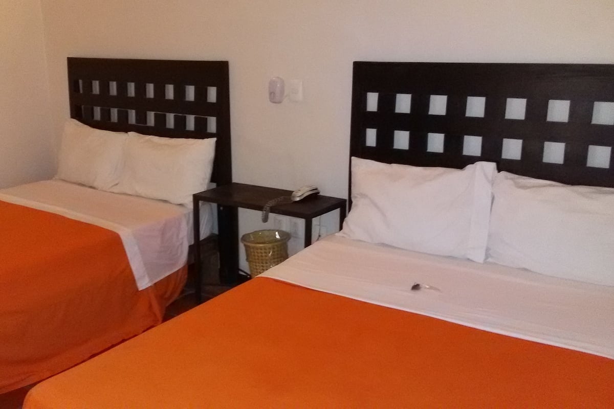 Twee eenpersoonsbedden met oranje beddengoed, een klein tafeltje met een telefoon en een prullenbak, en een lichtschakelaar aan de muur.