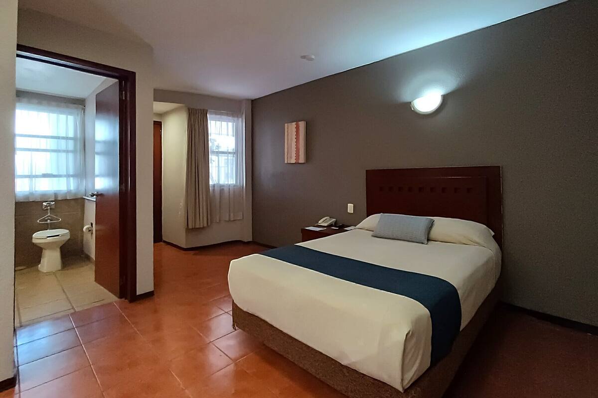 A hotel room with a bed, a bedside table, a nightstand, a window with curtains, and a view of the bathroom.